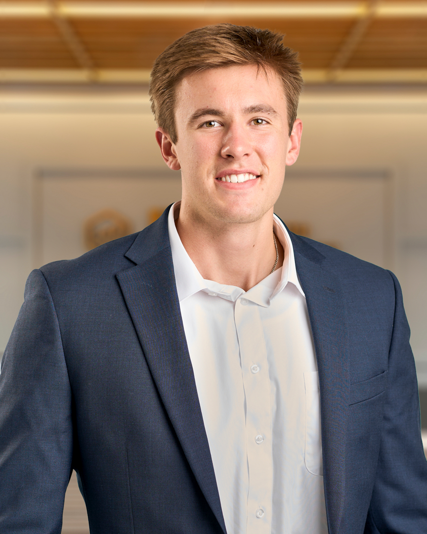 Adam, a man with short blonde hair smiling in a dark grey blazer over a white button up shirt, professional headshot with a warm lit office background.
