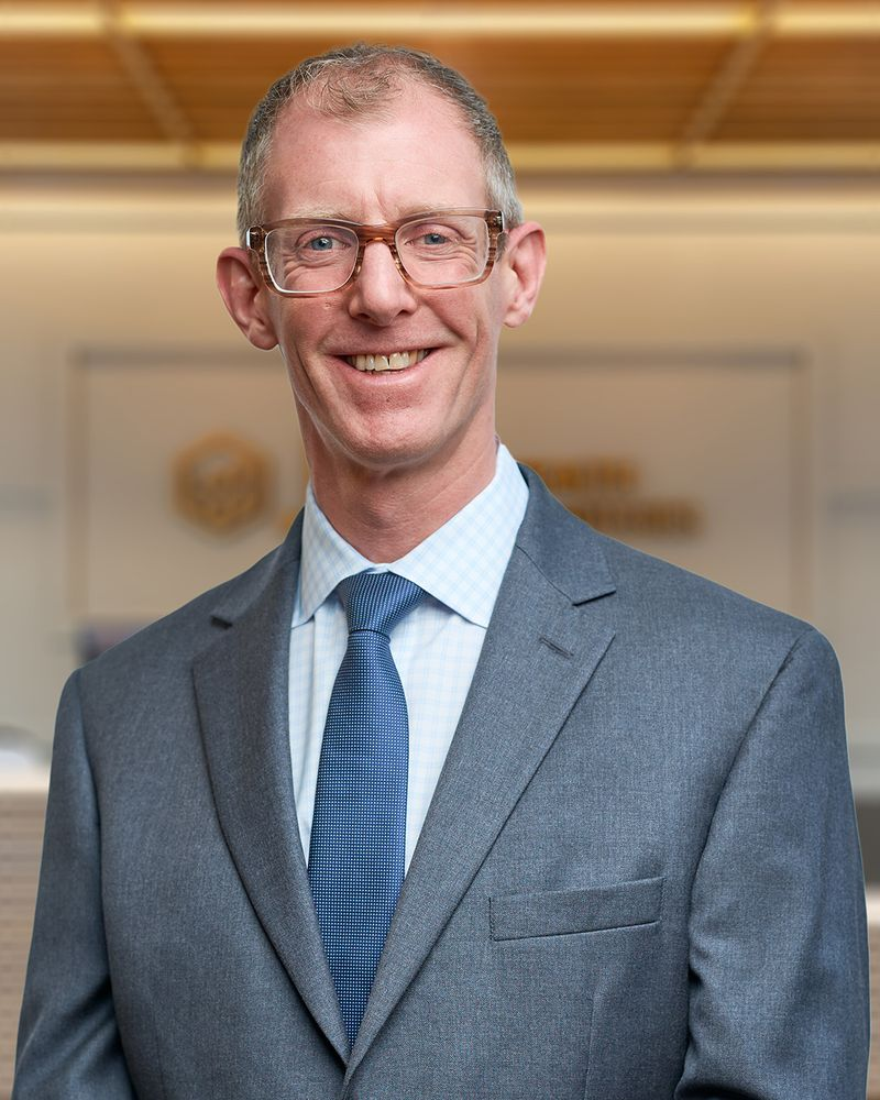 Adam, a man with short blonde hair smiling in a dark grey blazer over a white button up shirt, professional headshot with a warm lit office background.