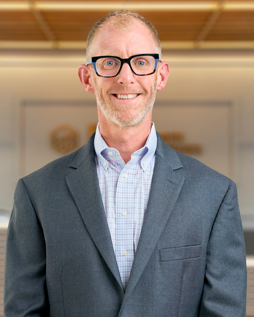 Adam, a man with short blonde hair smiling in a dark grey blazer over a white button up shirt, professional headshot with a warm lit office background.